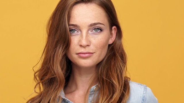 Close up of Carefree ginger woman in denim shirt corrects her appearance and looking at the camera over yellow background