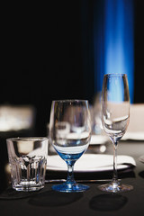Close up three glasses, forks, knives, napkins, candles and decorative flower on dark black tablecloth on table served for dinner in cozy restaurant. Empty served restaurant table with wine glass.