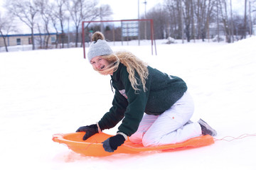 A laughing teenaged girl in ski pants and snow hat over her long hair sliding down a hill on a toboggan in a winter landscape