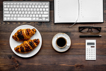 Morning of businessman. Coffee and croissants for breakfast near keyboard and notebook. Dark wooden background top view