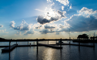 Fototapeta premium Dramatic sunset over a quiet marina: sunbeams pierce stormy clouds, silhouetted docks and a lone figure reflect on calm water—moody coastal scene.
