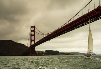 Fototapeta premium Moody view of the Golden Gate Bridge arching over choppy San Francisco Bay under overcast skies, a lone sailboat skimming the water near the bridge.