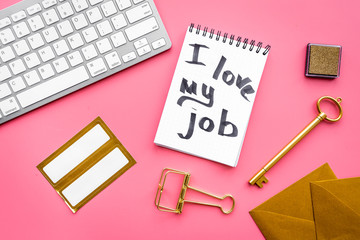 Happy at work. Handwritten motto I love my job in notebook near stationery on pink background top view