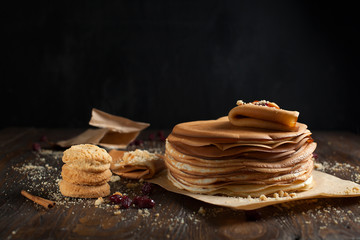 stack of pancakes, folded pancake, sprinkled with berries, cookies, sugar powder and caramel syrup on parchment on a dark wooden table