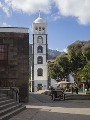 Naklejka na ściany i meble Canary islands Garachico main square with stairs horse carriage group of tourist walking siting on the banch and relaxing with white church tower trees hills and blue sky background
