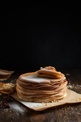 stack of pancakes, folded pancake, sprinkled with berries, cookies, sugar powder and caramel syrup on parchment on a dark wooden table