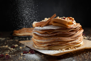 two stacks of pancakes, two folded pancakes, sprinkled with berries, cookies, sugar powder and caramel syrup on parchment on a dark wooden table