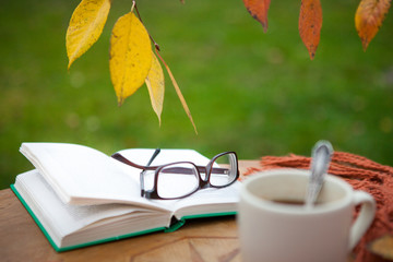 A cup of hot drink on the table in the garden. An open book with glasses. Plaid. Autumn.
