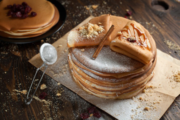 two stacks of pancakes, two folded pancakes, sprinkled with berries, cookies, sugar powder and caramel syrup on parchment on a dark wooden table