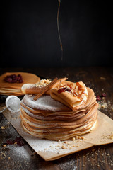 two stacks of pancakes, two folded pancakes, sprinkled with berries, cookies, sugar powder and caramel syrup on parchment on a dark wooden table