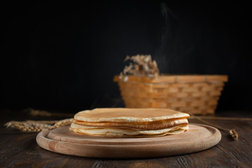 a pile of pancakes, spikelets, a round cutting board, a basket with branches with red berries and berries on a dark wooden table