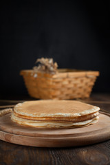 a pile of pancakes, spikelets, a round cutting board, a basket with branches with red berries and berries on a dark wooden table