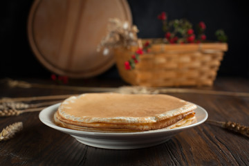 a pile of pancakes, spikelets, a round cutting board, a basket with branches with red berries and berries on a dark wooden table