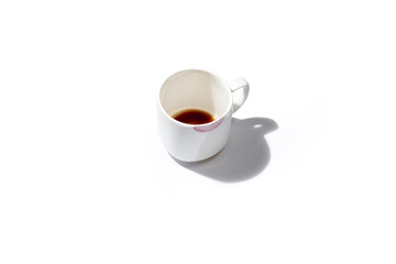 una tazzina bianca lasciata come gesto  di presenza quotidiana sul tavolo con fondo di caffè e sporca di rossetto sul bordo su fondo bianco e ombra di appoggio