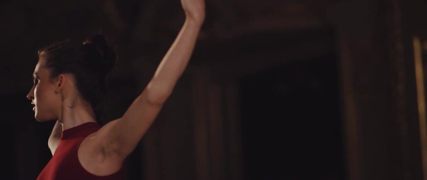 Professional ballerina dressed in red dress with open back dancing ballet with pointes in spotlights on big stage. Beautiful young girl performing with premiere.