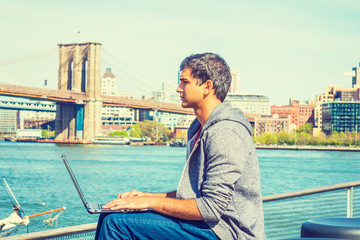 Fototapeta na wymiar Way to Success. East Indian American student travels, studies in New York, wearing hooded sweatshirt, jeans, sits by river, works on laptop computer, seriously thinks. Brooklyn bridge on background..