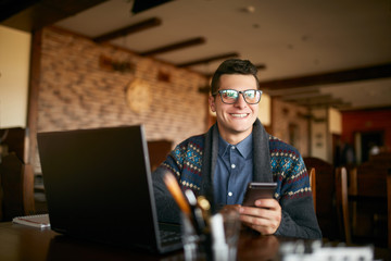 Successful businessman holding mobile modern smartphone. Happy hipster with a modern phone enjoys pleasant news. Smiling young man in glasses with laptop on table. Multitasking theme.