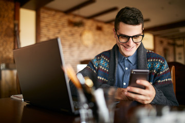 A young man typing a text on mobile modern smartphone. Hipster holding a modern phone and writing a phone message. Smiling young businessman in glasses looking at cellphone with laptop on table.