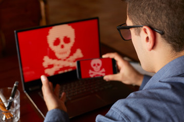 Man sits near laptop with phone blocked and encrypted by ransomware spyware asking for money....