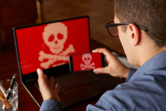 Man sits near laptop with phone blocked and encrypted by ransomware spyware asking for money. Laptop and smartphone infected by virus. Scary red skull crossbones on screen. Cyber security concept.
