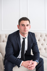 Portrait of a young serious and confident man in a suit sitting on the couch and looking at camera.