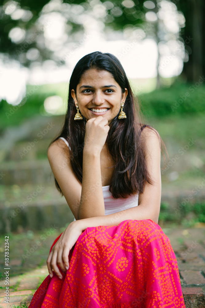 Wall mural Portrait of young Indian woman smiling and sitting in a park (Fort Canning Park in Singapore) in the day. She is attractive and is dressed in a summer outfit.