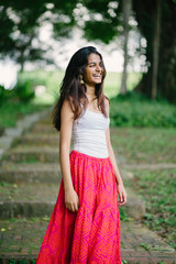 Candid portrait of a young and attractive Indian Asian woman in a lush, green park (Fort Canning Park in Singapore) on a sunny day. She is dressed in a fashionable, casual summer outfit. 