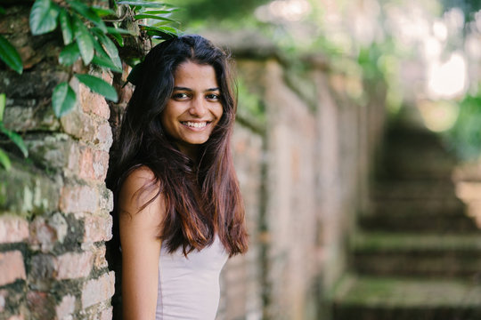 Candid Portrait Of A Young And Attractive Indian Asian Woman In A Lush, Green Park (Fort Canning Park In Singapore) On A Sunny Day. She Is Dressed In A Casual Summer Outfit And Leaning Against A Wall.