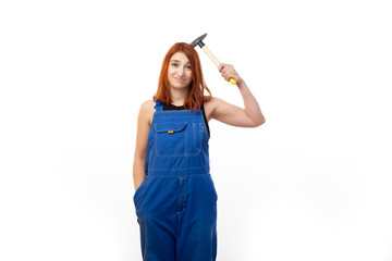  A young red-haired woman in a black T-shirt and blue construction overall smiles and funny keeps a hammer over his head on a white isolated background