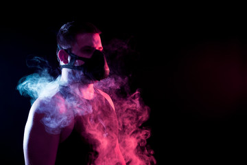 A dark-haired man breathes through a training black mask around a blue and red cloud of smoke from a wipe on a black isolated background