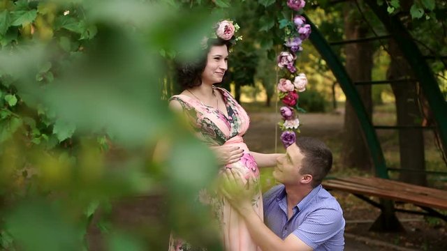 Husband tenderly hugging pregnant wife. Husband caressing wife's belly. A pregnant woman in a beautiful long dress with flowers in the Park with her husband.