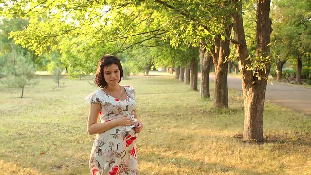 Portrait of happy pregnant girl in summer park at sunset. Beautiful pregnant brunette in a forest in summer, the wind is waving her dress.