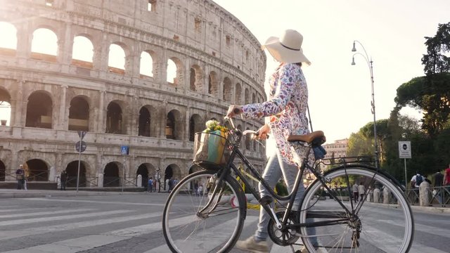 Beautiful young woman in colorful fashion dress walking alone with bike crossing road in front of colosseum in Rome at sunset happy attractive tourist girl with straw hat
