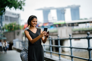 Candid portrait of a young Indian Asian woman (tourist) looking at her smartphone with the Singapore city skyline in the back. 