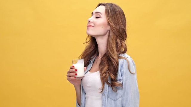 Happy brunette woman in denim shirt drinking kefir and looking at the camera over yellow background