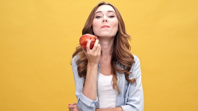 Cheerful brunette woman in denim shirt eating big fresh apple over yellow background