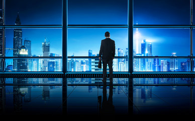 Fototapeta na wymiar Back view of thoughtful businessman looking out of big office window at office ,planning future project concept, night scene .
