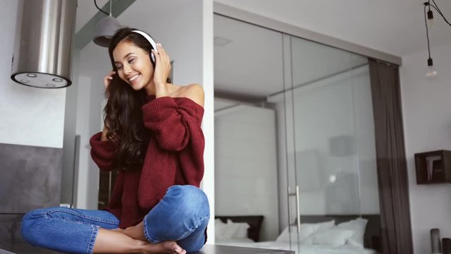 Happy brunette woman in sweater sitting on table at home and listening music