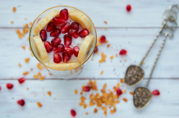Yogurt with fresh sliced banana and pomegranate seeds on the white wooden table. Vintage spoons with chia, scattered granola grains and pomegranate seeds on the blurred background. Top view