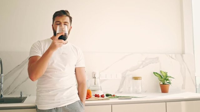 Smiling bearded man drinking wine on kitchen and looking away
