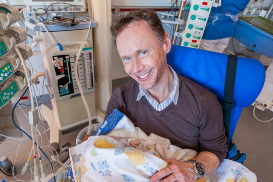 A father sitting on a chair in a intensive care unit holding his sick infant boy wrapped in a blanket surrounded by medical equipment.