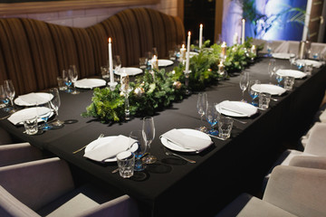 Glasses, forks, knives, napkins, candles and decorative flower on dark black tablecloth on table served for dinner in cozy restaurant. Empty served restaurant table with plates and wine glass.