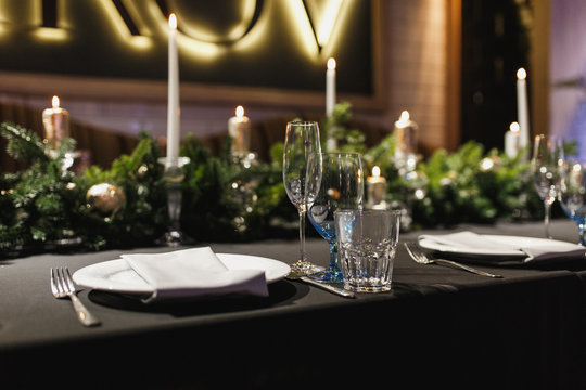 Close up glasses, forks, knives, napkins, candles and decorative flower on dark black tablecloth on table served for dinner in cozy restaurant. Empty served restaurant table with plates and wine glass