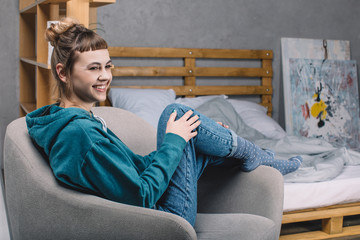 smiling girl sitting on armchair in bedroom and looking away