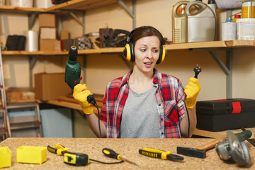Perplexed young woman in plaid shirt gray T-shirt noise insulated headphones yellow gloves working in carpentry workshop at wooden table place with piece of wood, different tools, power drill, wire.