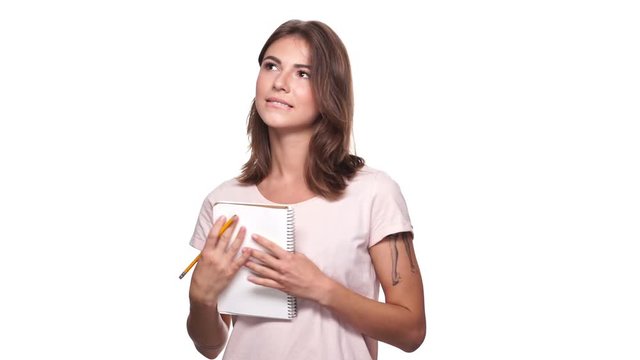 Pleased pensive brunette woman in t-shirt writing something in notebook over white background