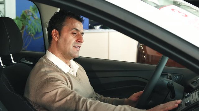 A man is sitting in the car. He holds his hands on the steering wheel and presses the buttons. Then he looks at the camera and smiles