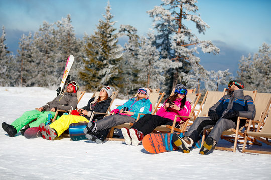 friends sitting with deck chairs in winter mountains. Sunbathing in snow