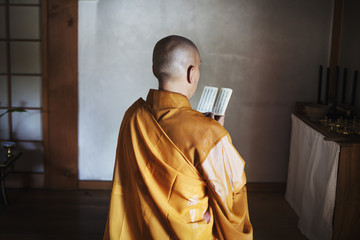 Rear view of Buddhist monk with shaved head wearing golden robe sitting indoors in a temple, holding prayer text.