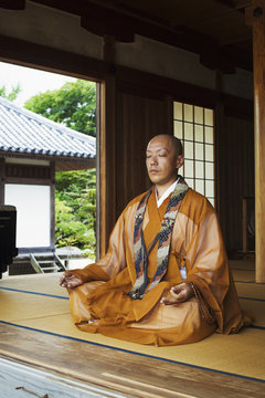 Buddhist monk with shaved head wearing golden robe sitting cross legged on the floor, meditating, Buddhist hand gesture.